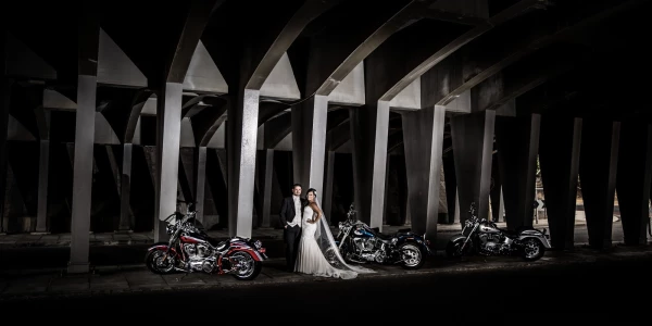 Our couple, Yaira + Sean had their wedding in early January on a super hot day, The heat was intense and there was no breeze to lessen the temperature effect. This did not stop our couple from enjoying every single minute of their wedding day though. The groom’s father brought 3 brand new Harley Davidson motorbikes especially for the wedding day, they were driven as an escort ahead of the wedding cars. So, we had to come up with a great photo idea to incorporate them in. <br />
<br />
The entire wedding party was in love with the idea of doing a shot under this old bridge in Fremantle, WA. They got some much needed shade from the sun’s intense heat; and the low light + concrete walls and columns were a great background to create this shot.<br />
<br />
We used 4 off camera flashes to illuminate the couple and the bikes so that we could darken everything else right down.<br />
<br />
This image is now a double page spread in their wedding album.